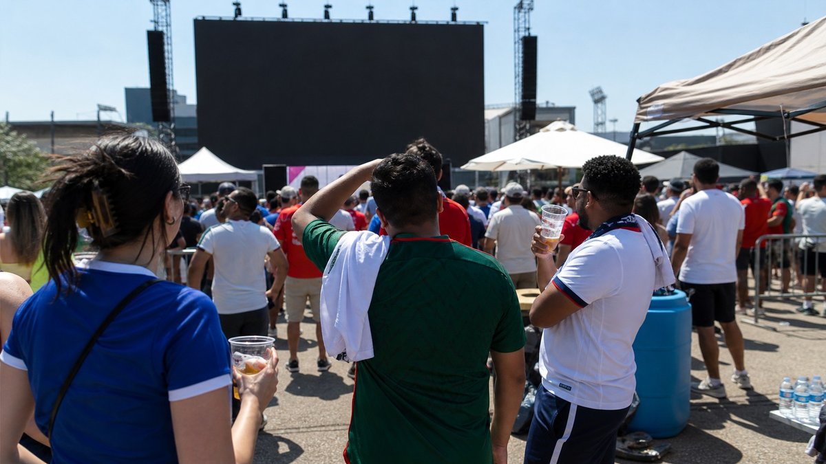 Torcedores de futebol em fan zone sob sol forte com copos e garrafas de água.